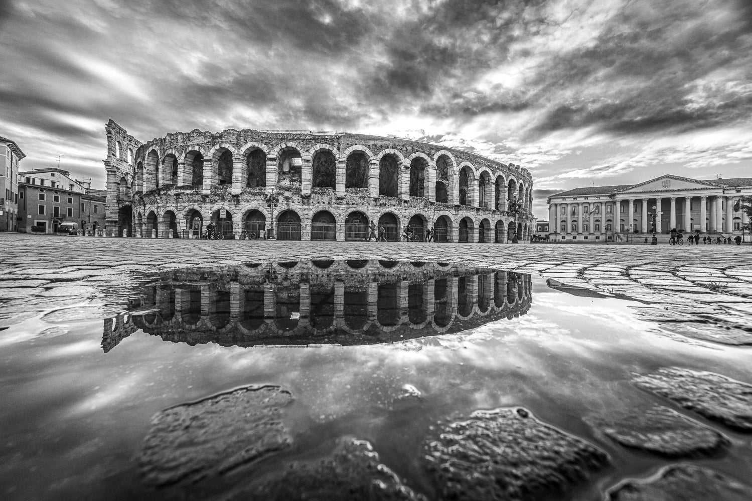 Fotografia Fine Art in bianco e nero Verona Collection #08645 con Arena di Verona riflessa in una pozzanghera su Piazza Bra, cielo nuvoloso e luce diffusa.