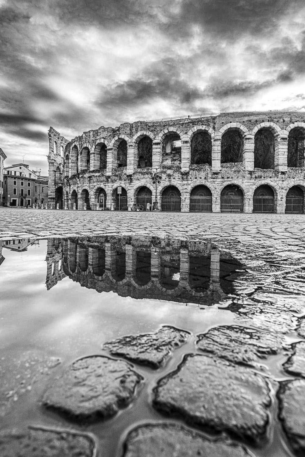 Fotografia Fine Art in bianco e nero verticale Verona Collection #08654 con l’Arena di Verona riflessa sull’acqua dopo un temporale. Il selciato in primo piano e il cielo drammatico esaltano la composizione architettonica.